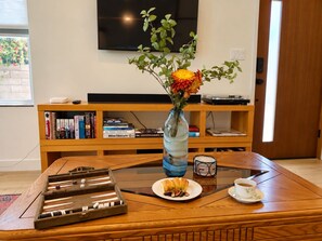 Un salon avec une table basse, un jeu de société, une assiette de pâtisseries, un vase de fleurs, une bibliothèque et un téléviseur.
