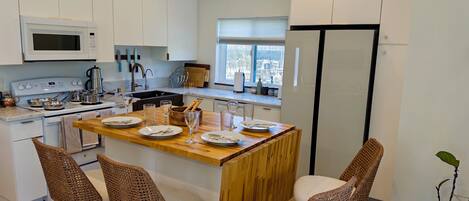 Una cocina moderna con armarios blancos, una isla de madera con taburetes de bar, un refrigerador de acero inoxidable y un ventanal que proporciona luz natural.