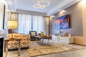 A modern living room with a sofa, a coffee table, a TV mounted on the wall, and a large window with curtains. - 3-bedroom apartment in Westlands perfect Family friendly retreat, Pool, Gym, Wow (Nairobi)