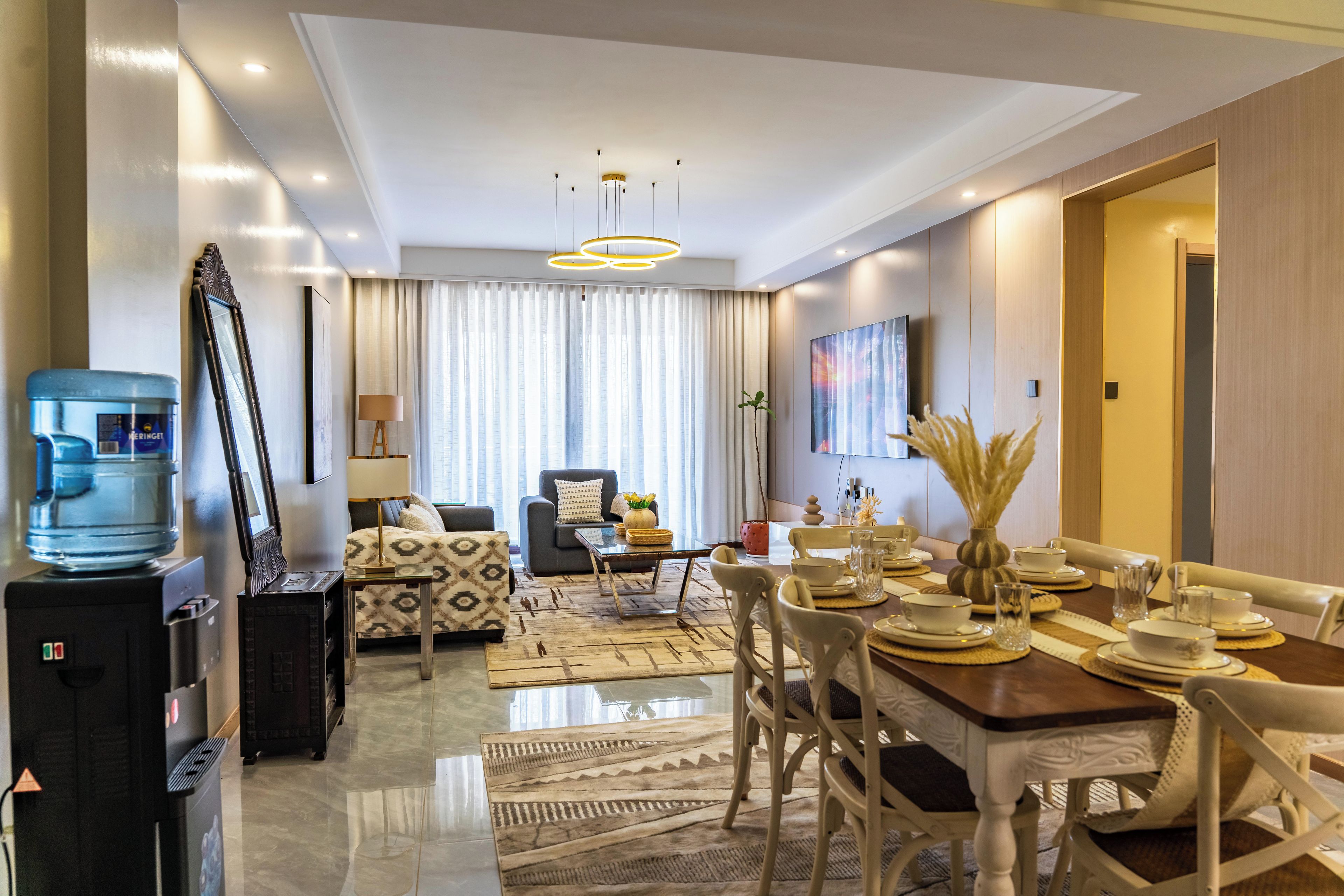 A modern living room with a dining area, a water dispenser, and a large window with curtains.