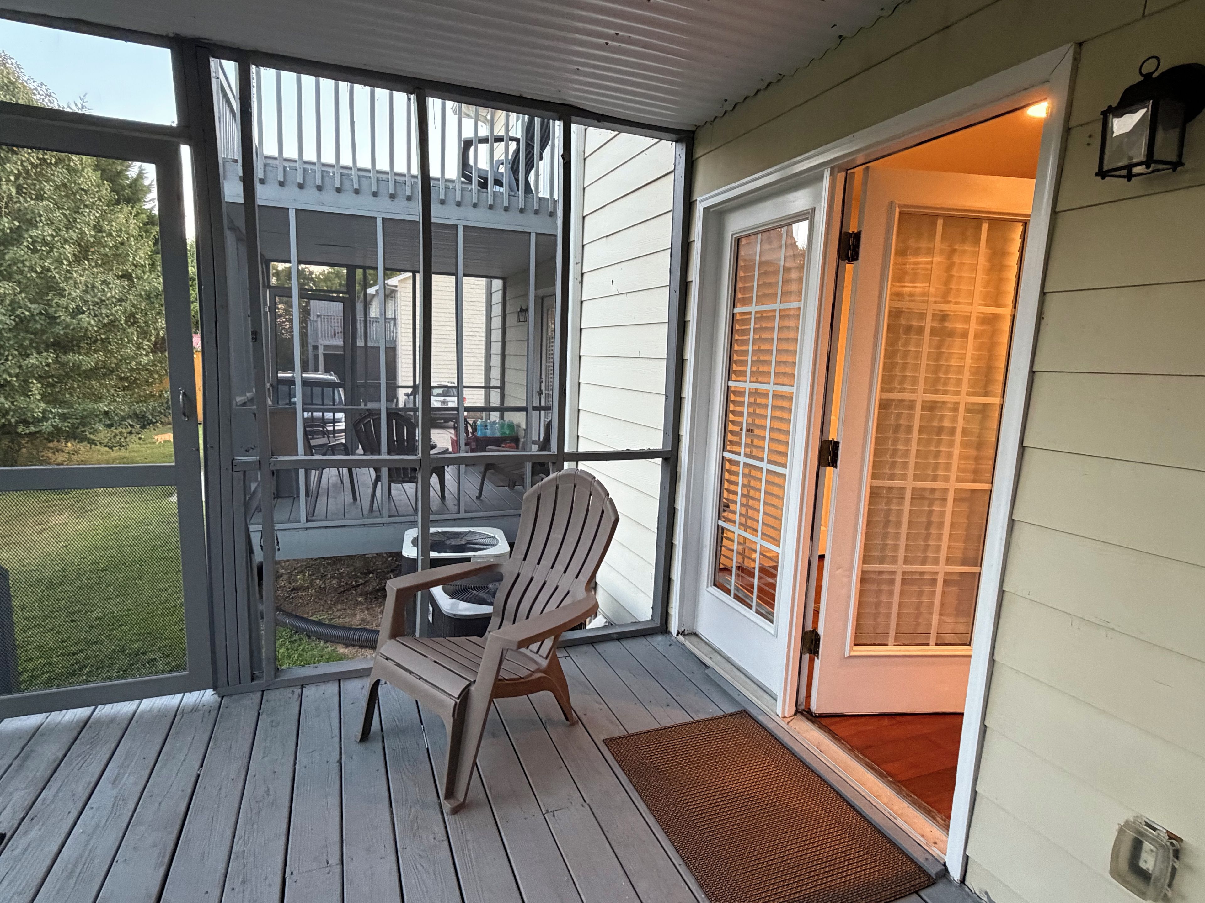 Une véranda couverte avec une chaise en bois, une table et une vue sur le jardin à travers une porte moustiquaire.