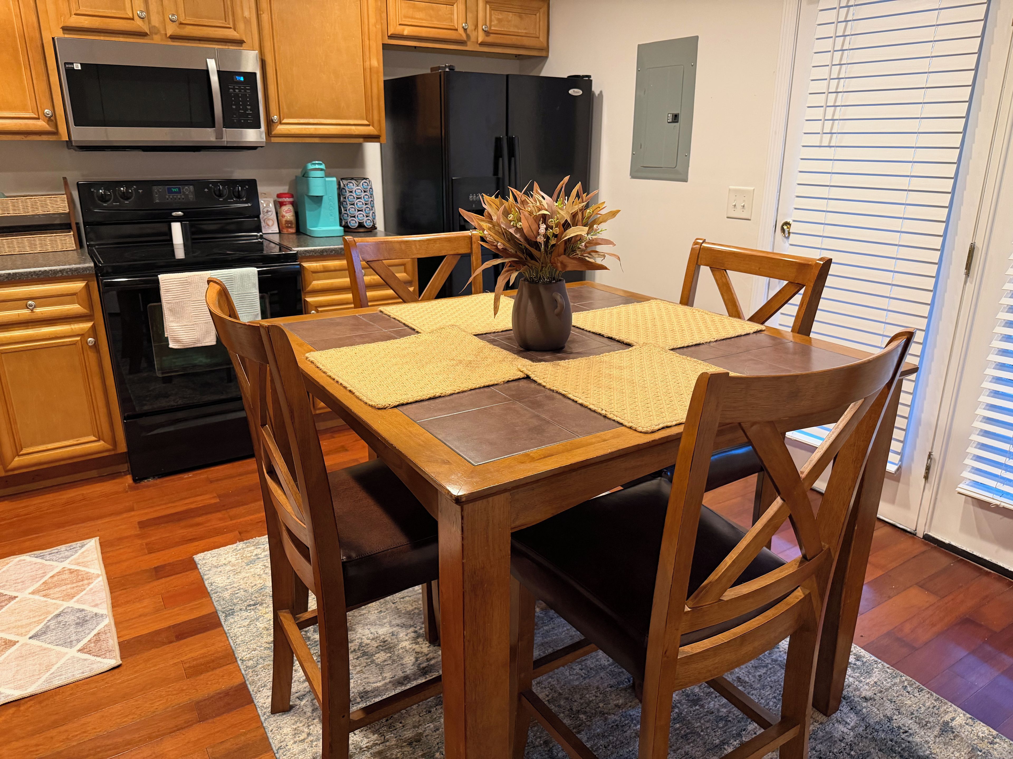 A modern kitchen with wooden cabinets, a black refrigerator, a microwave, and an oven. There is a dining table with a vase of flowers and placemats.