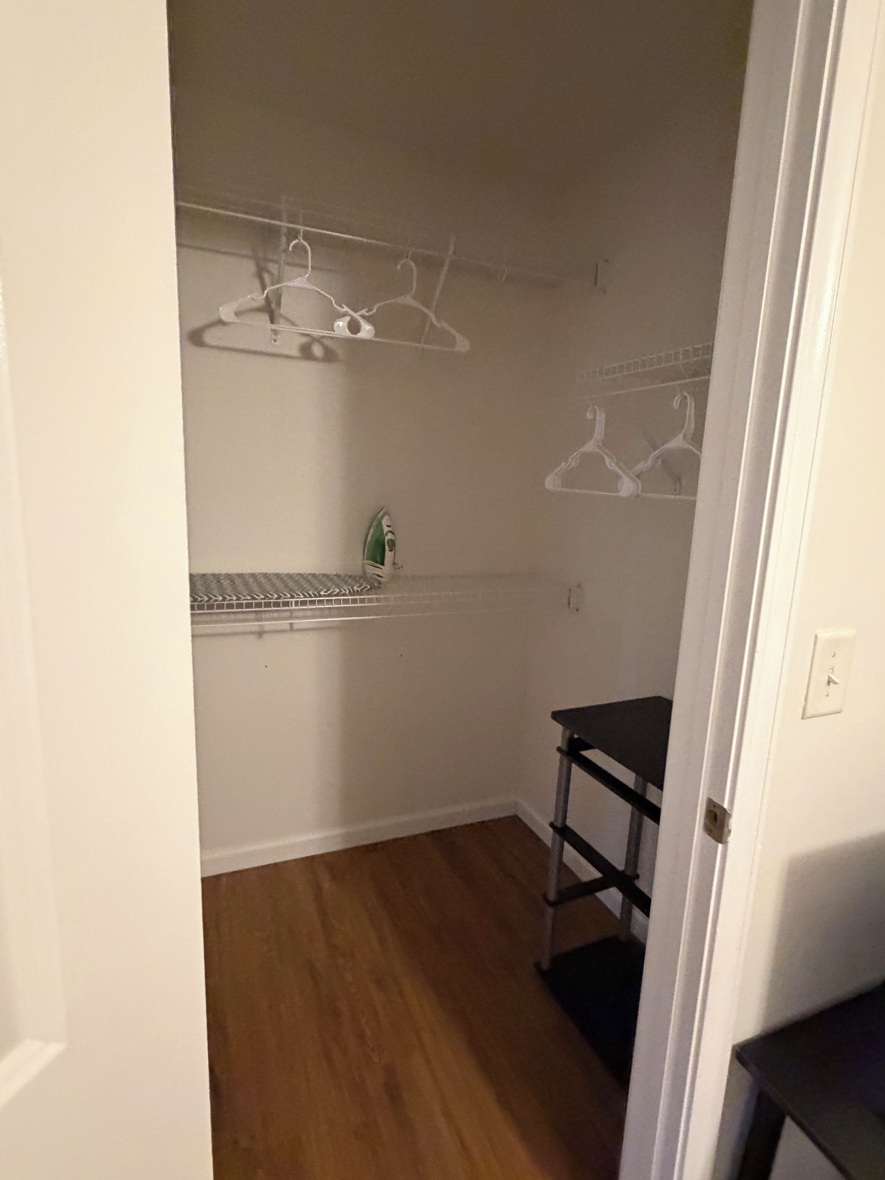 Um quarto pequeno e estreito com piso de madeira, uma prateleira de metal com roupas penduradas e um pequeno ferro de passar.