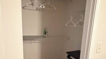 Um quarto pequeno e estreito com piso de madeira, uma prateleira de metal com roupas penduradas e um pequeno ferro de passar.