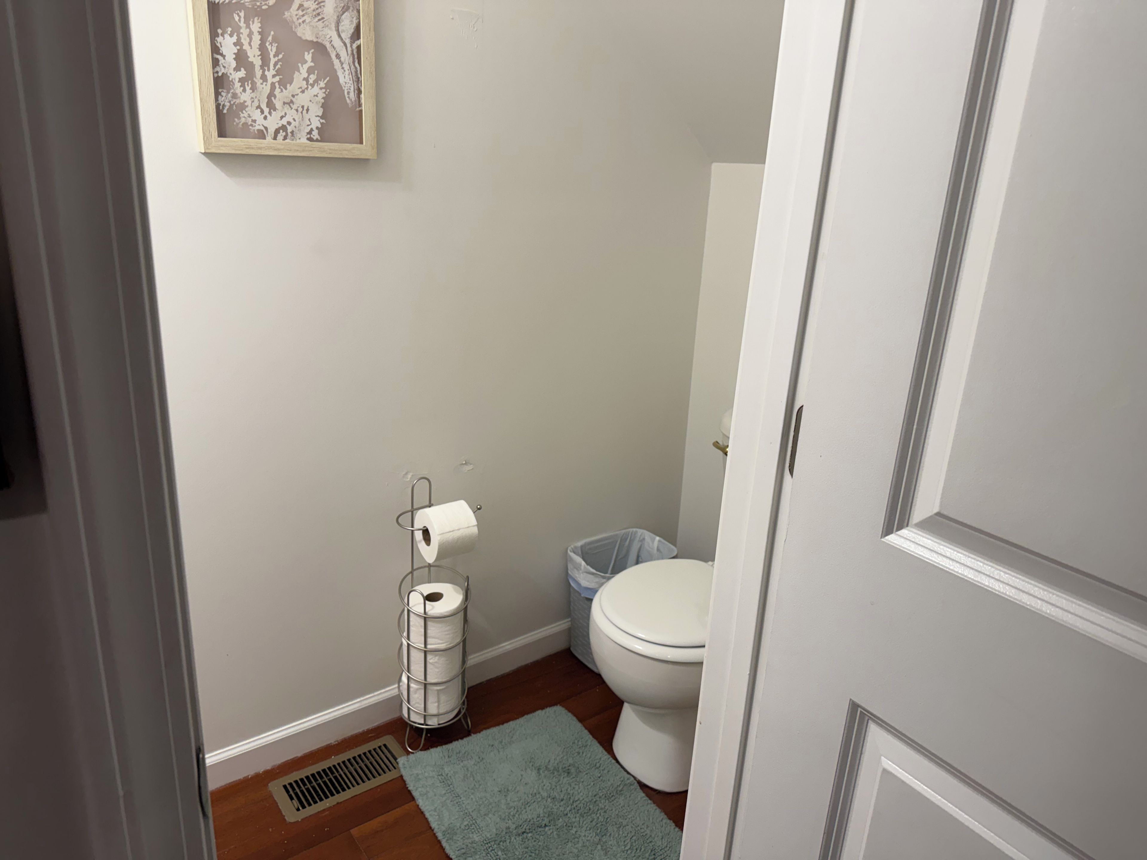 A bathroom with a toilet, a roll of toilet paper, a trash bin, a vent, and a framed picture on the wall.