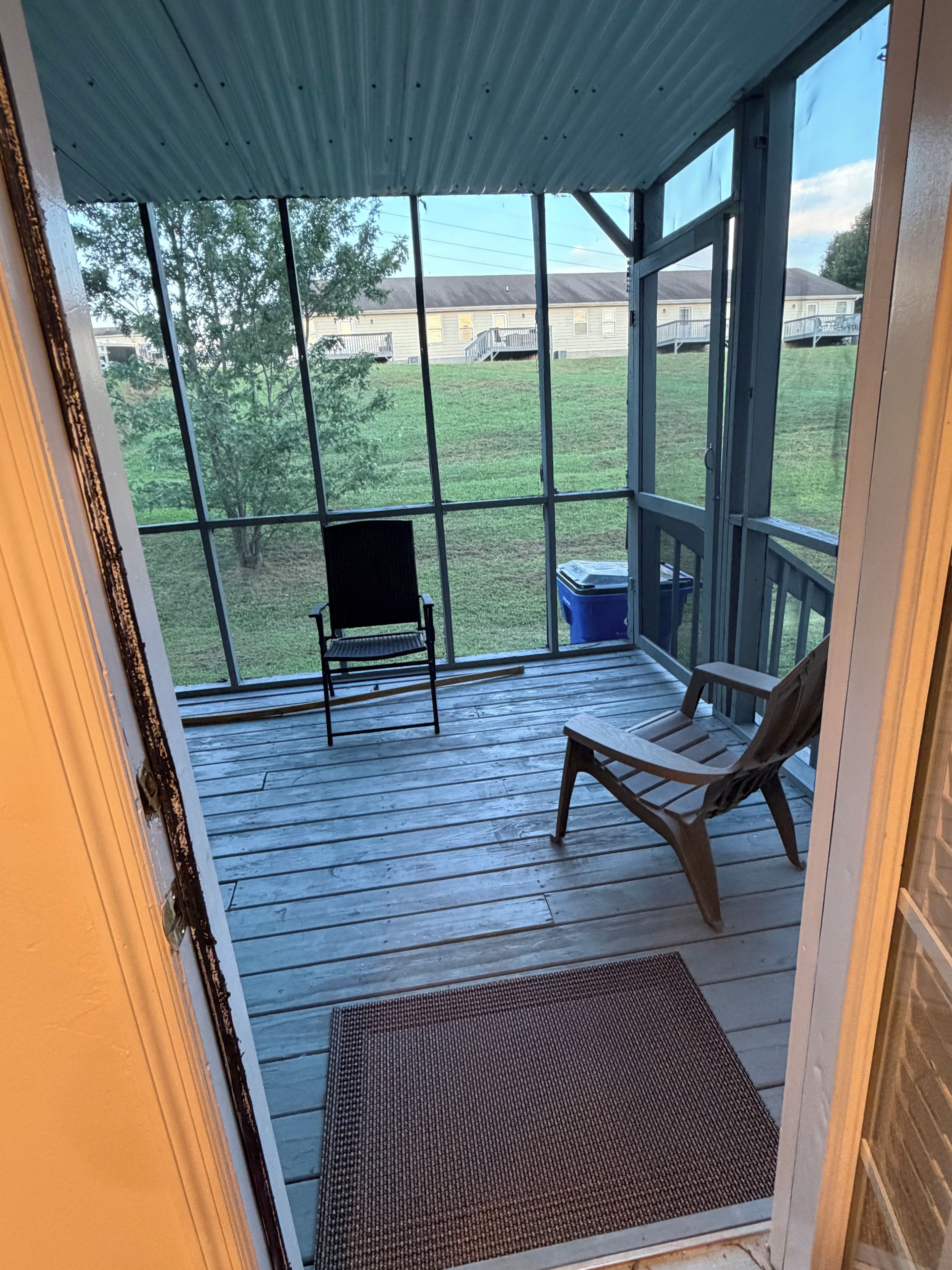A covered porch with a wooden floor, a bench, a chair, and a view of a grassy yard and buildings in the distance.