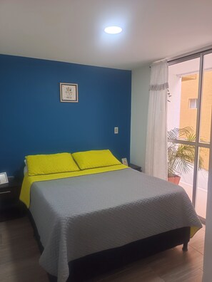 Ein Schlafzimmer mit einem Bett, einem Nachttisch, einer Wandleuchte, einem Fenster mit Vorhängen und Blick nach draußen.