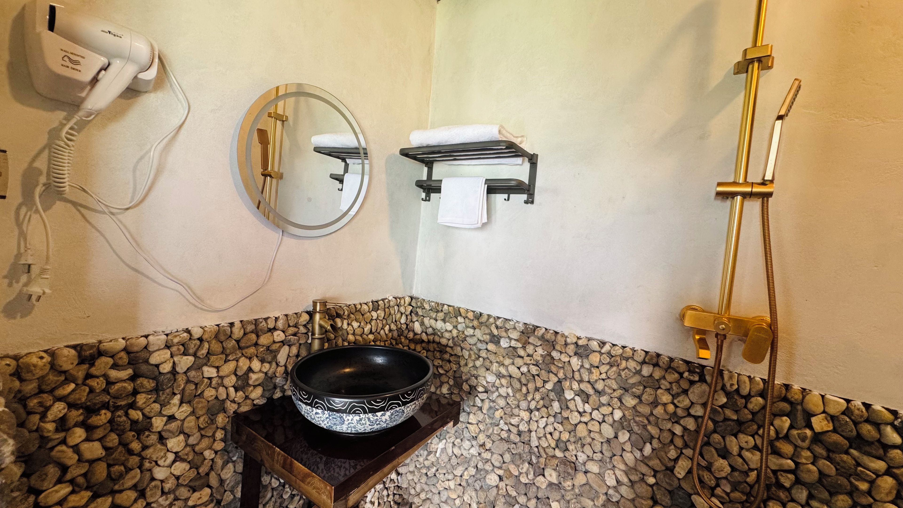Un baño con suelo de baldosas de piedra, un lavabo negro sobre un soporte de madera, un secador de pelo, un espejo y un perchero para toallas.