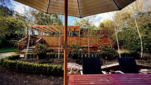 A wooden deck with a staircase leading to a covered area, surrounded by potted plants and trees.