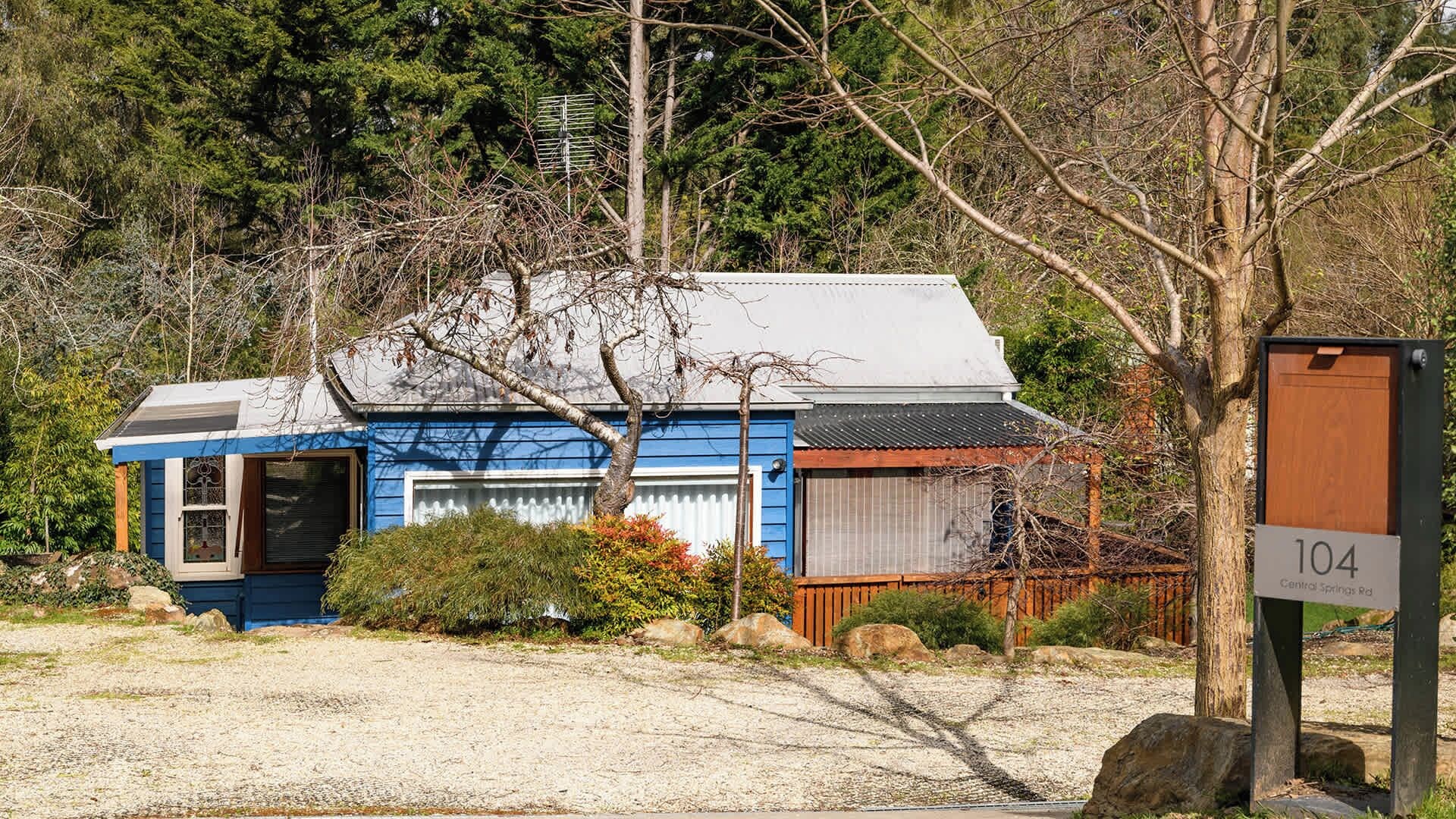 A small, single-story building with a blue exterior and a grey roof, surrounded by trees and a gravel driveway.