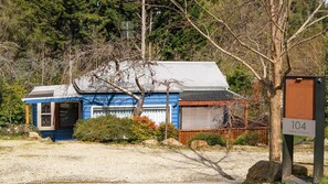A small, single-story building with a blue exterior and a grey roof, surrounded by trees and a gravel driveway.
