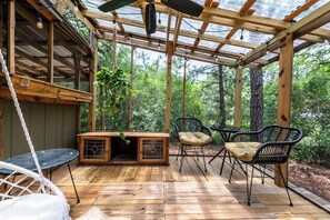 Una terraza de madera con techo de vidrio, rodeada de árboles, con una silla colgante, una mesa y dos sillas.