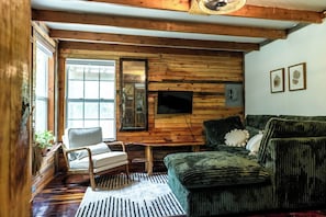 Interior acogedor de una cabaña con un muro de madera, un sofá verde, una silla blanca y un televisor montado en la pared.