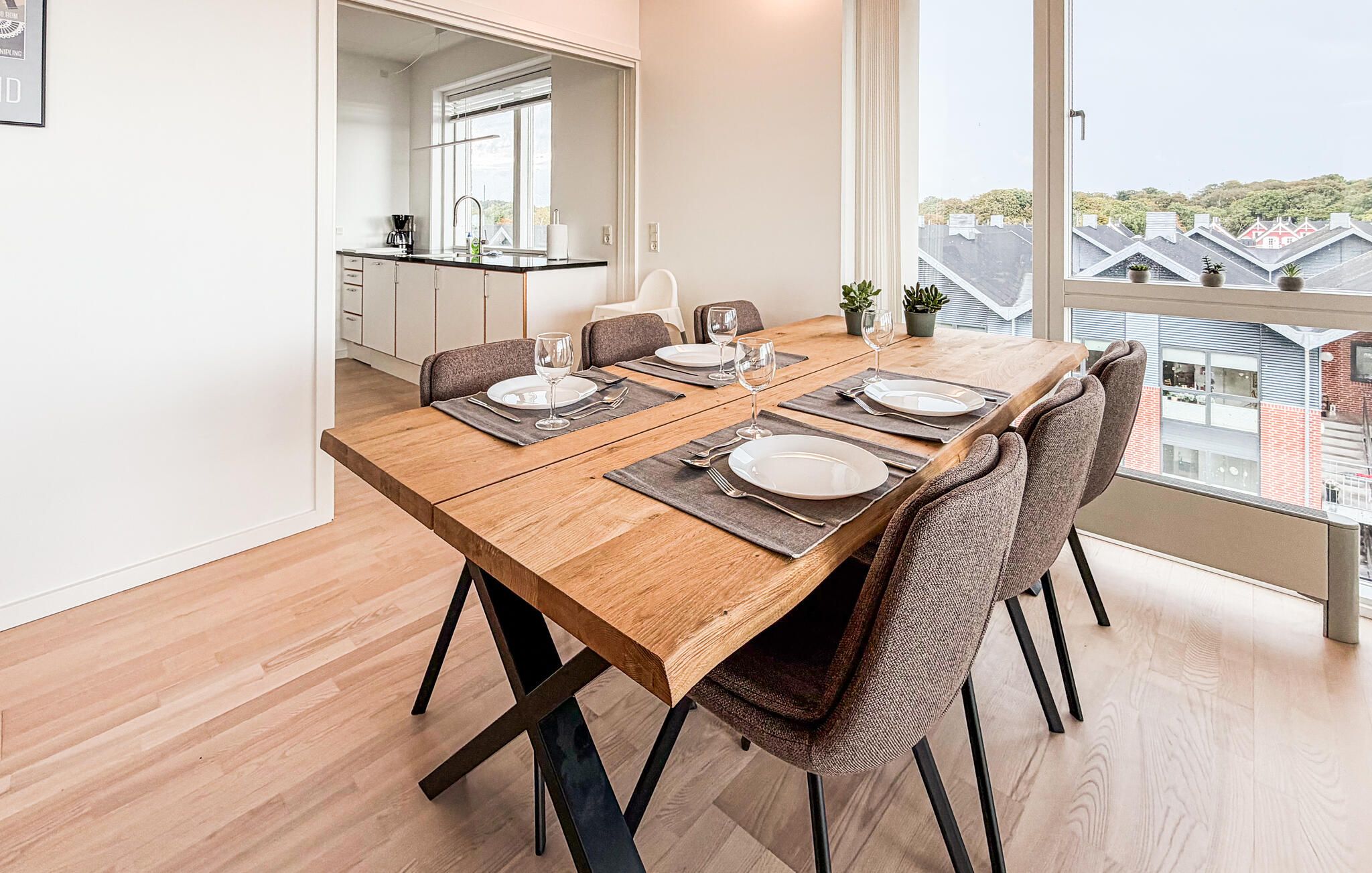 A dining area with a wooden table set for four, surrounded by chairs, in a room with a large window offering a view of buildings.