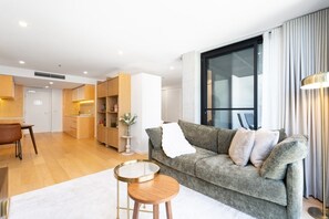 A modern living room with a grey sofa, a round wooden coffee table, and a dining area with a brown chair. - High-End 1BR with Enclosed Balcony Near ANU (Braddon)