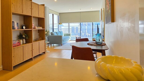 Een moderne woonkamer met een houten boekenkast, een eettafel met stoelen, een bank en een groot raam met uitzicht op de stad.