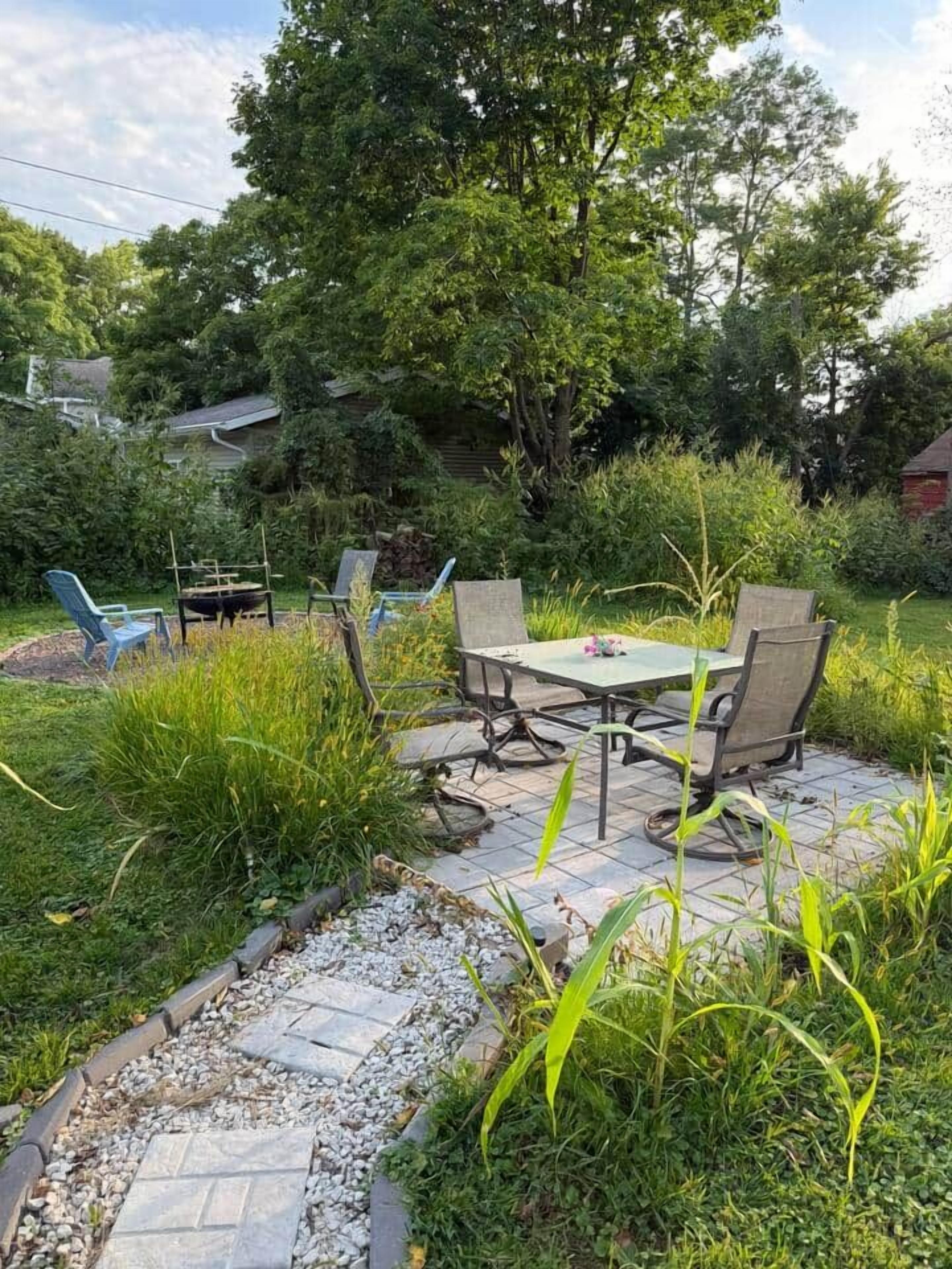 Une terrasse extérieure avec une table et des chaises, entourée de verdure et bordée d’un chemin en pierre.