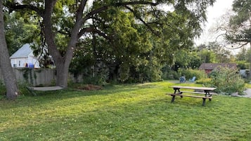 A grassy outdoor area with a picnic table and a bench, surrounded by trees and a fence.