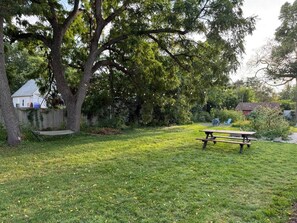 Un espacio al aire libre con césped, una mesa de picnic y un banco, rodeado de árboles y una cerca.