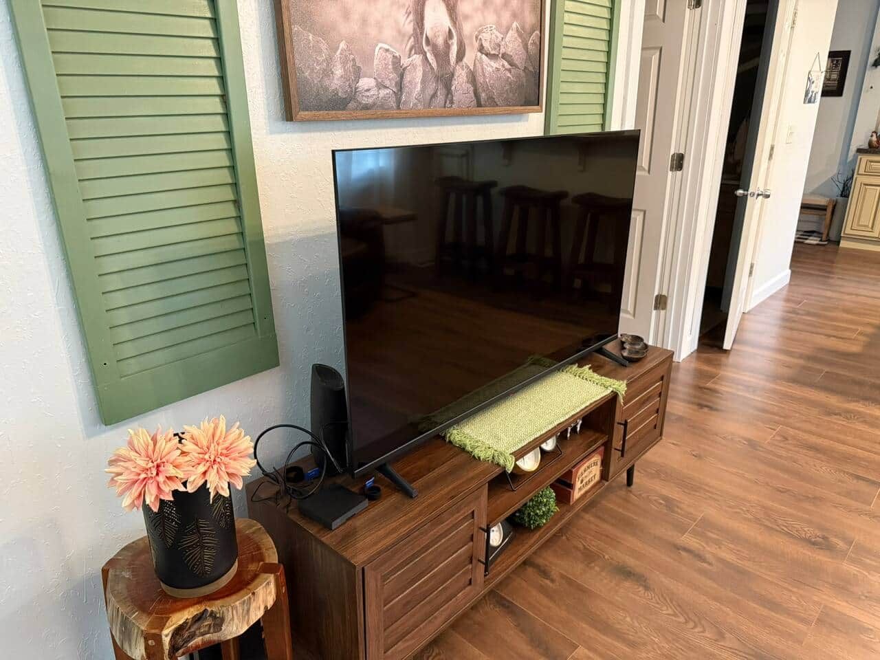 A flat-screen TV mounted on a wooden entertainment center with a green throw blanket and a vase of flowers.