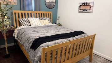 A bed with a wooden frame, a grey quilt, and a black throw blanket. There is a plant to the left and a wall-mounted picture frame to the right.