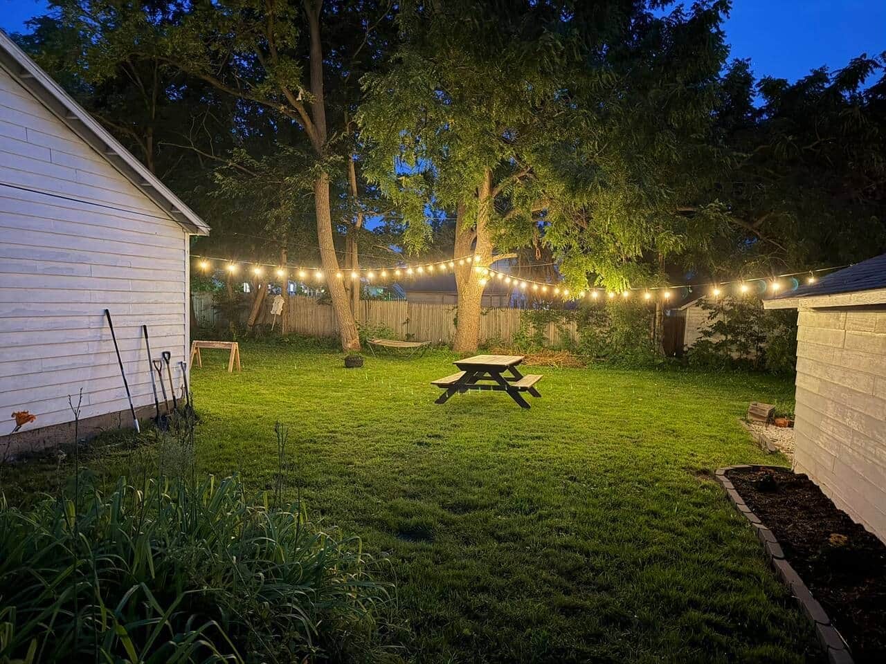 Un jardin avec une table de pique-nique, des guirlandes lumineuses et une clôture en bois.