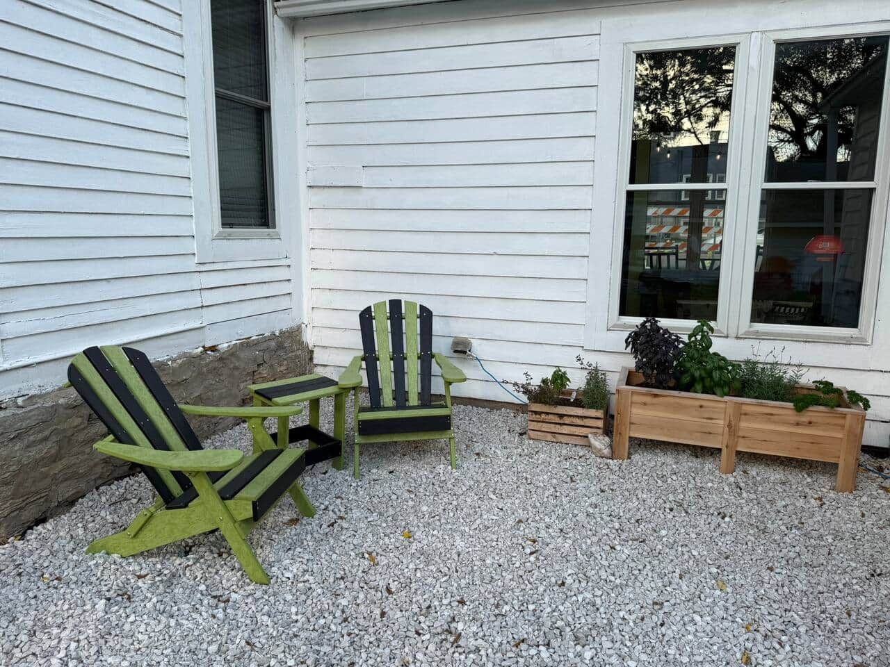 Een terras met Adirondack-stoelen en een kleine tafel, een houten plantenbak met planten, en een gebouw met ramen.