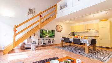 A modern living room with a wooden staircase, a dining area with a table set for two, and a kitchen in the background.