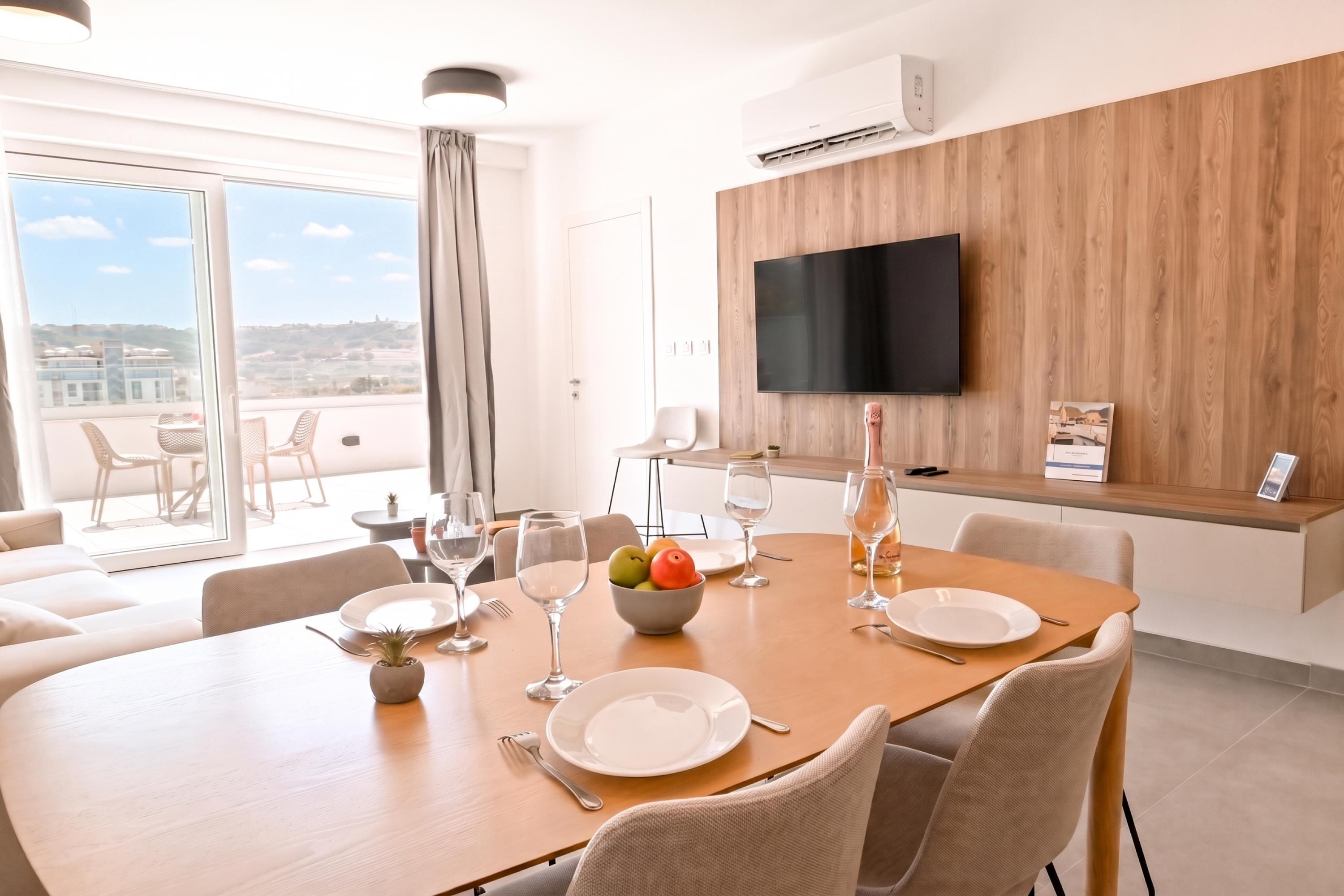 Un espace repas moderne avec une grande table dressée pour quatre personnes, un téléviseur à écran plat fixé à un mur en bois et un balcon avec des sièges d’extérieur.