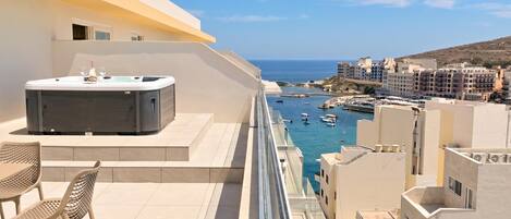 A balcony with a hot tub, chairs, and a view of the sea and buildings.