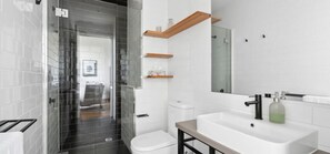 A modern bathroom with a white sink, a mirror, and a toilet. There are wooden shelves above the sink and a glass shower door. - Devlin 103 - Stylish Geelong Apartment (Greater Geelong)