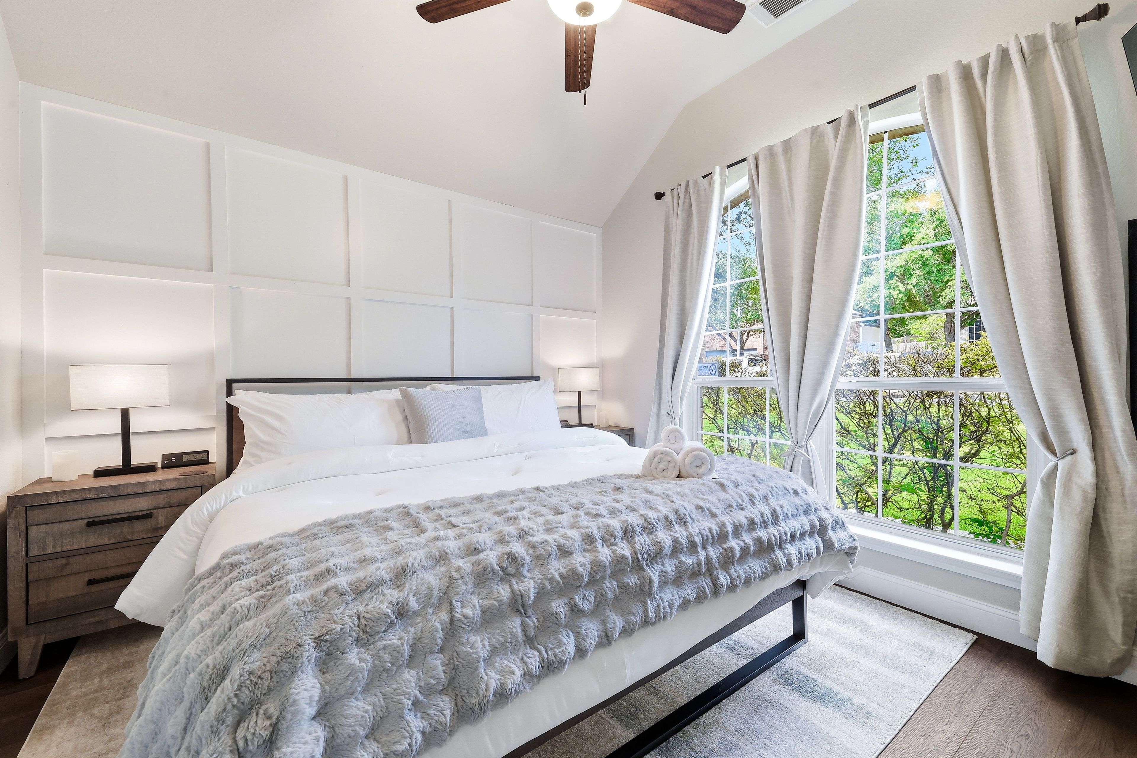 Ein Schlafzimmer mit einem großen Bett, Nachttischen, einem Deckenventilator und einem Fenster mit Vorhängen.