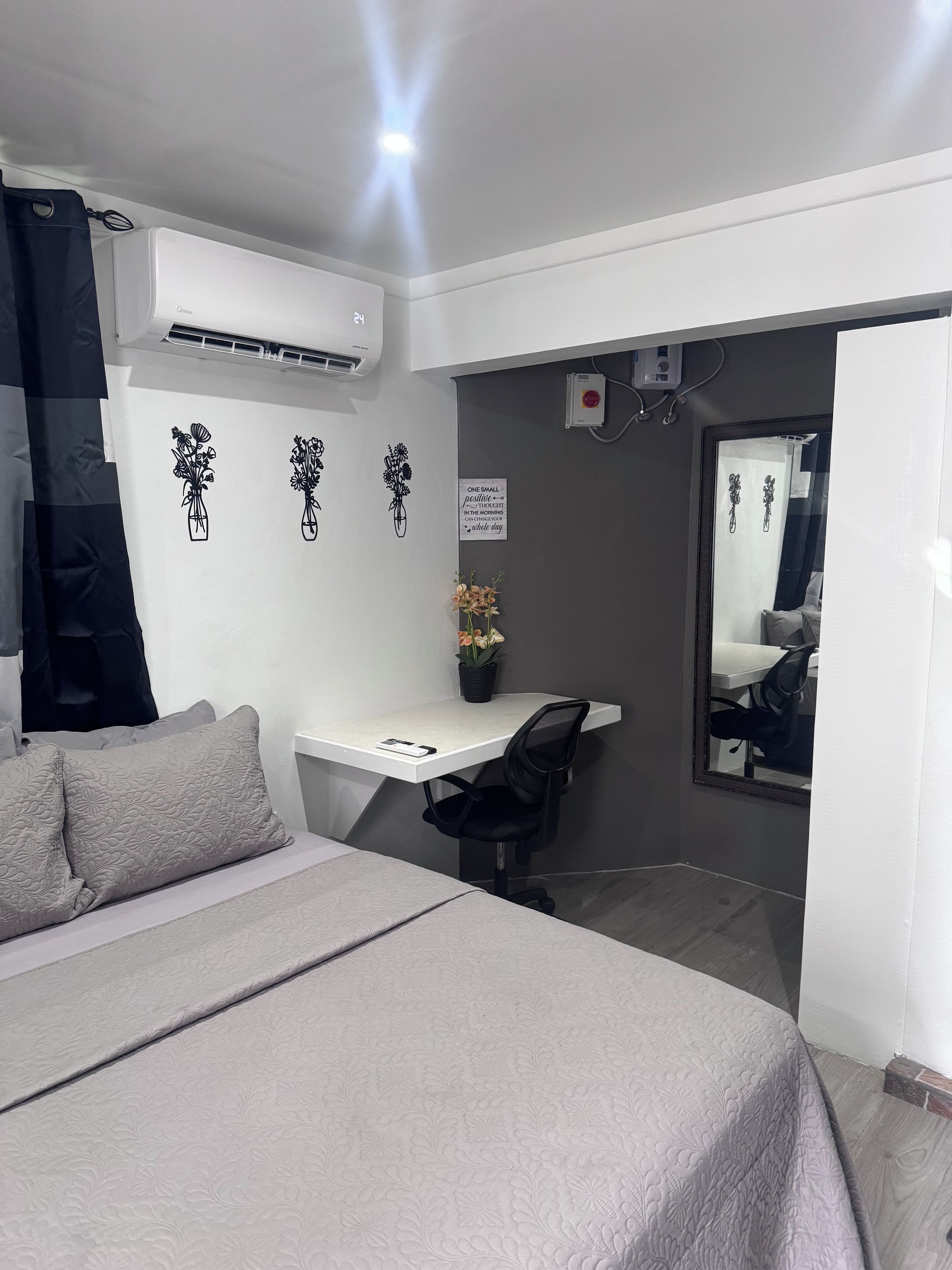 Une chambre avec un lit, un bureau et une chaise. Un climatiseur est fixé au mur et une petite plante en pot est posée sur le bureau.