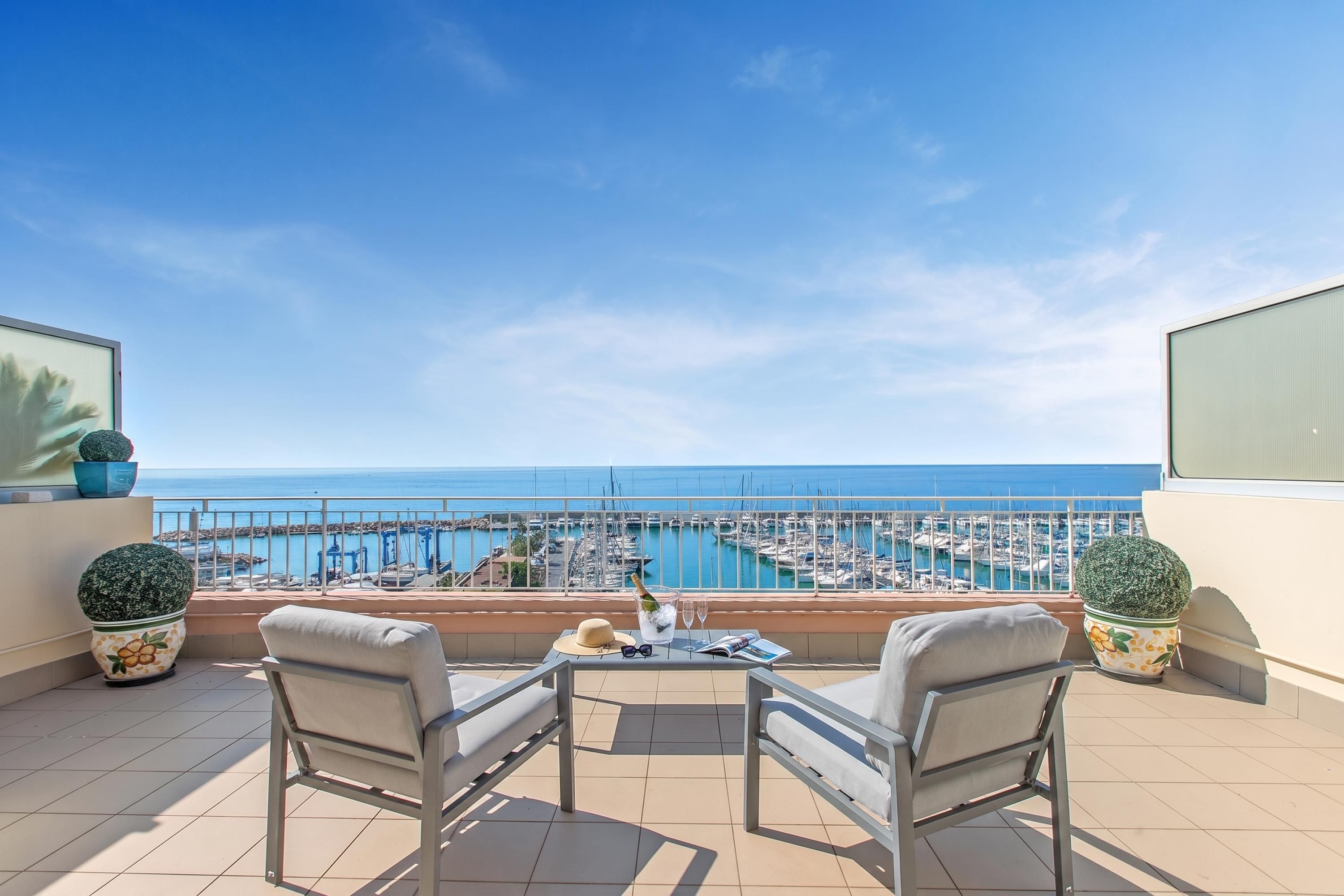 A balcony with two chairs, a table, and a view of a marina and sea.