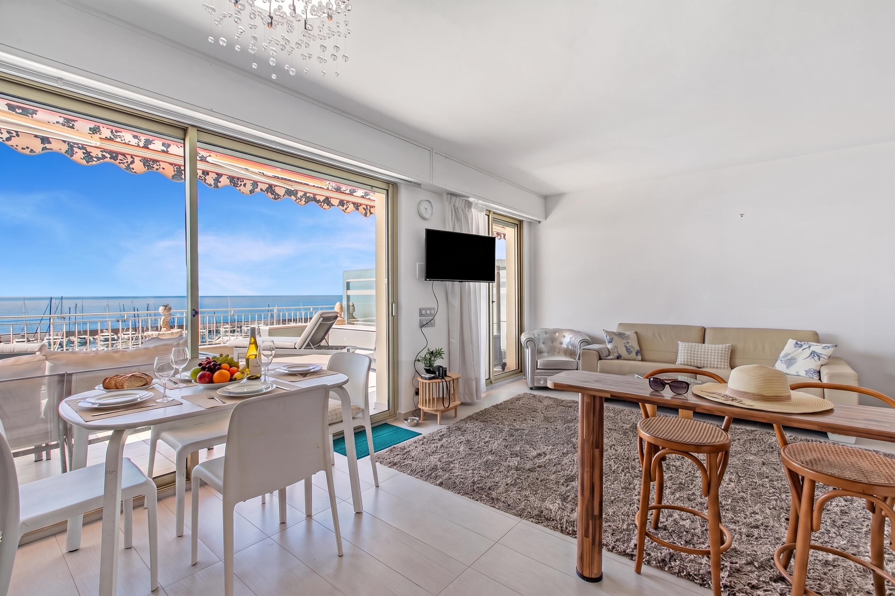 A modern living room with a large window overlooking a marina, a dining area with a fruit bowl and wine bottle, and a cozy seating area with a TV.
