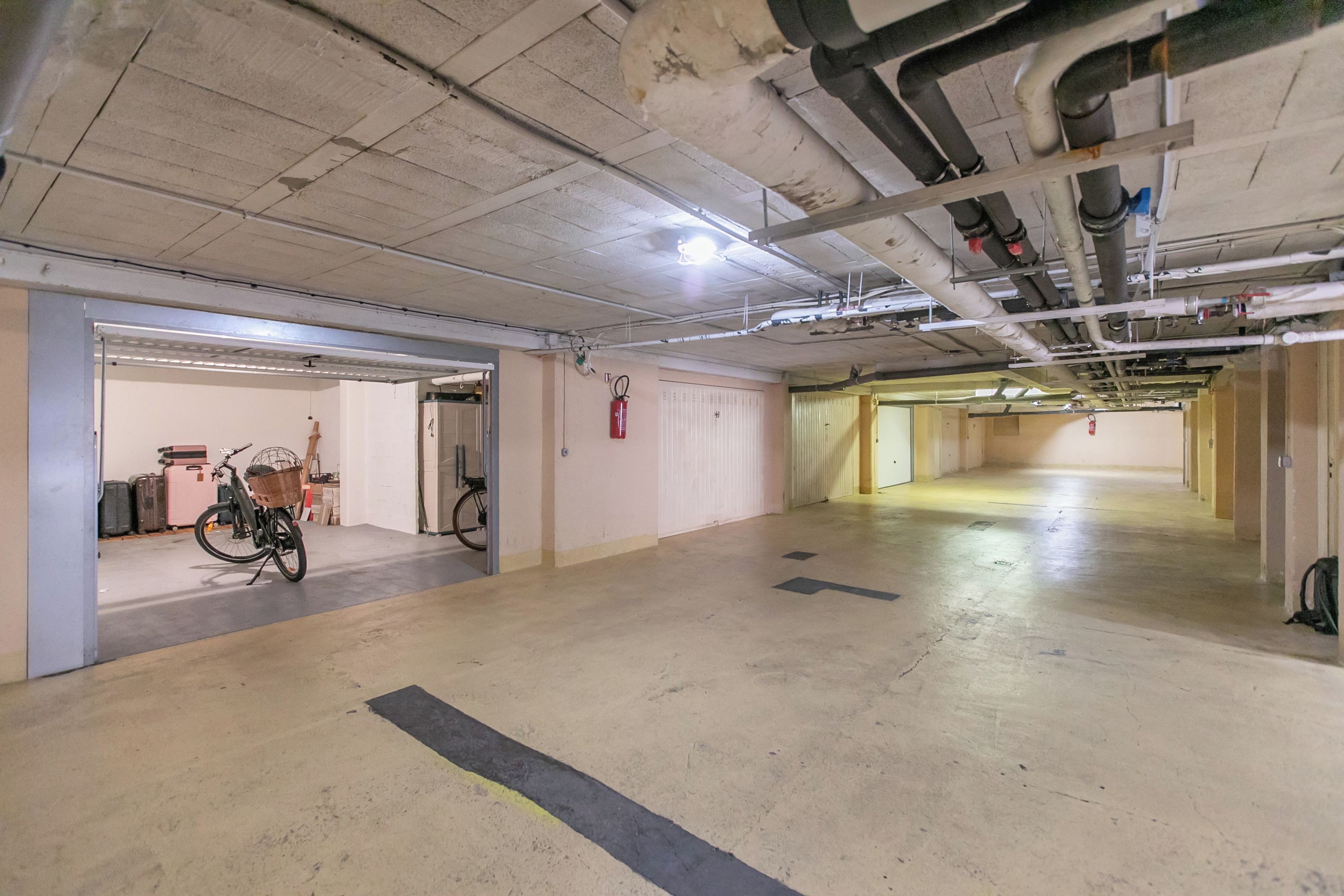 Un vaste espace de stationnement souterrain, doté de sols et de murs en béton, d’un extincteur fixé au mur et d’un vélo garé dans un coin.