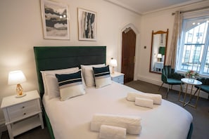 A hotel room with a large bed, two bedside tables, a green upholstered chair, a small round table, and a window with curtains.
