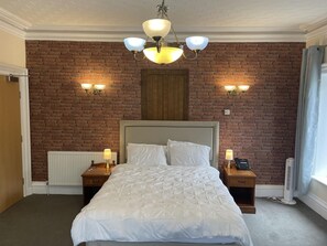 A hotel room with a brick wall, a large bed with white bedding, two bedside tables with lamps, a wooden headboard, and a ceiling light fixture.