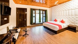 A hotel room with a wooden ceiling, a large bed with red pillows, a TV mounted on the wall, and a window with green curtains.