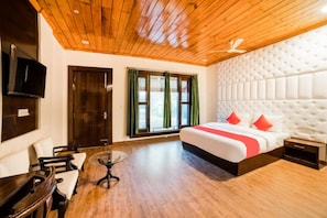 A hotel room with a wooden ceiling, a large bed with red pillows, a TV mounted on the wall, and a window with green curtains.
