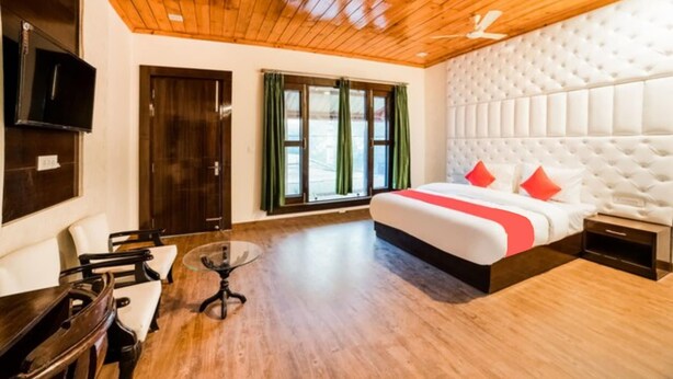 A hotel room with a wooden ceiling, a large bed with red pillows, a TV mounted on the wall, and a window with green curtains. - Goroomgo Blue Moon Bhimtal (Nainital)
