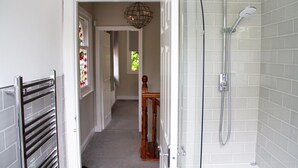 A bathroom with a shower, a towel rack, and a door leading to a hallway with a wooden chair and a stained glass window.