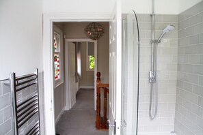 A bathroom with a shower, a towel rack, and a door leading to a hallway with a wooden chair and a stained glass window.