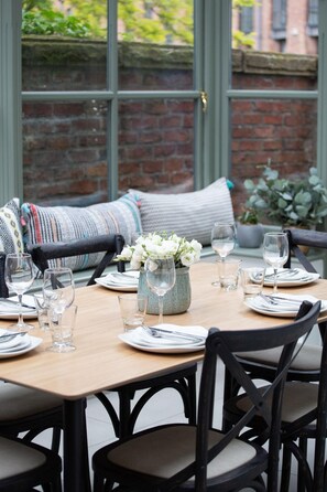 A dining table set for six with chairs, a vase of white flowers, and a brick wall in the background.