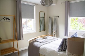 A bedroom with a bed, a bedside table, a mirror, a window with curtains, a hanging rack, and a lamp. - Lock Keepers Cottage - Detached House in the City (Manchester)
