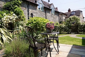 A stone house with a garden, two metal chairs, and a table with a flower pot.