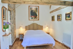 Un dormitorio con cabecera de madera, cama con ropa de cama blanca, dos lámparas de noche, un espejo, un florero con flores y un cuadro enmarcado en la pared.