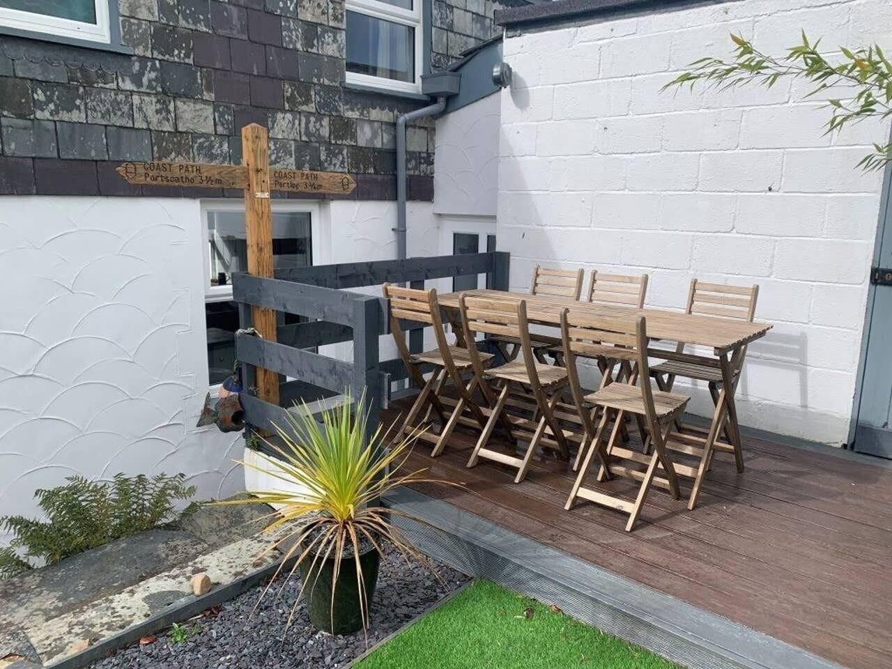 A wooden table and chairs set up on a patio with a white wall and a wooden fence in the background.