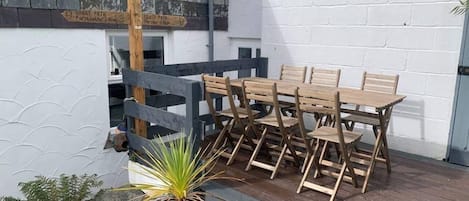 A wooden table and chairs set up on a patio with a white wall and a wooden fence in the background.