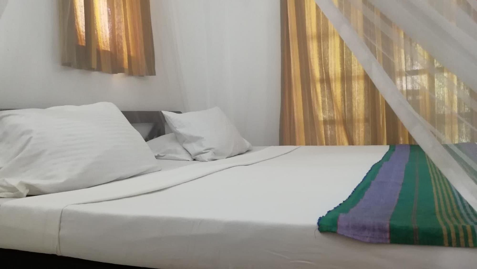 Una cama con ropa de cama blanca y una manta a rayas, una mesita de noche y una ventana con cortinas.